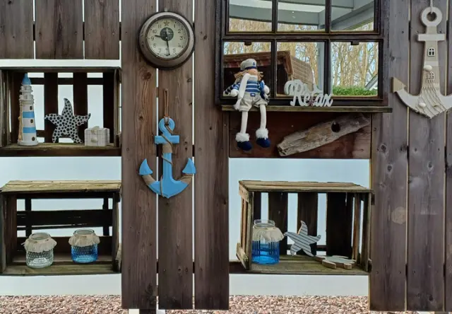 Eine rustikale Holzwand zeigt nautisches Dekor, darunter eine Uhr, einen Anker, Regale, Seesterne und eine Matrosenpuppe vor einer gemütlichen Kulisse.