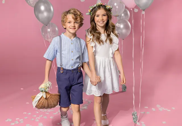 Zwei Kinder halten sich an den Händen. Die Kinder tragen festliche Klamotten und der Hintergrund ist rosa