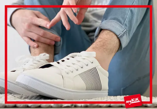 Eine Person in lässigen weißen Turnschuhen rückt ihr Hosenbündchen zurecht und präsentiert einen entspannten Stil auf einer strukturierten Oberfläche.